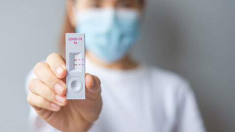 Scharf im Vordergrund ist ein positiver Corona-Selbsttest zu sehen, den eine Frau in die Kamera hält. Sie ist nur unscharf im Hintergrund zu sehen und trägt eine OP-Maske und ein weißes T-Shirt.