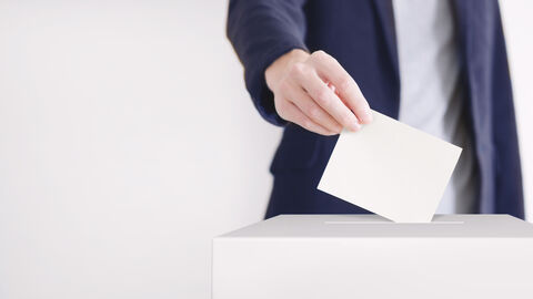 Hand mit Stimmzettel, der in Wahlurne geworfen wird.