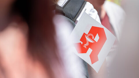 Nahaufnahme von einem Fähnchen. Darauf ist das rote Zeichen der Apotheke auf weißen Untergrund.