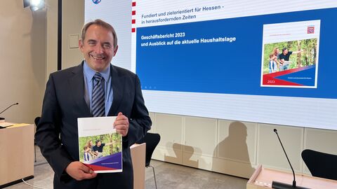 Zu sehen ist Hessens Finanzminister Professor Dr. R. Alexander Lorz im Presseraum des Hessischen Landtags, er hält den Geschäftsbericht 2023 in seinen Händen. 