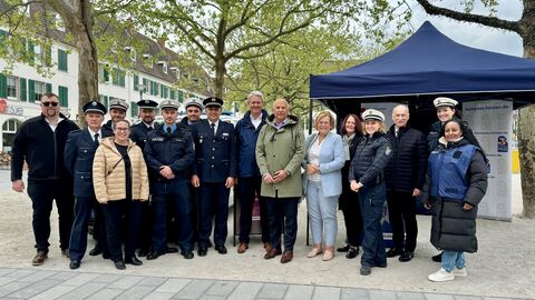 Innenminister Roman Poseck (mitte) Oberbürgermeister Patrick Burghardt (links), Landtagsabgeordnete Sabine Bächle-Scholz (rechts), Landespolizeipräsident Robert Schäfer, Polizeipräsident Björn Gutzeit mit Mitarbeiterinnen und Mitarbeitern der Polizeistation Rüsselsheim gemeinsam auf einem Platz vor einem Zelt. 
